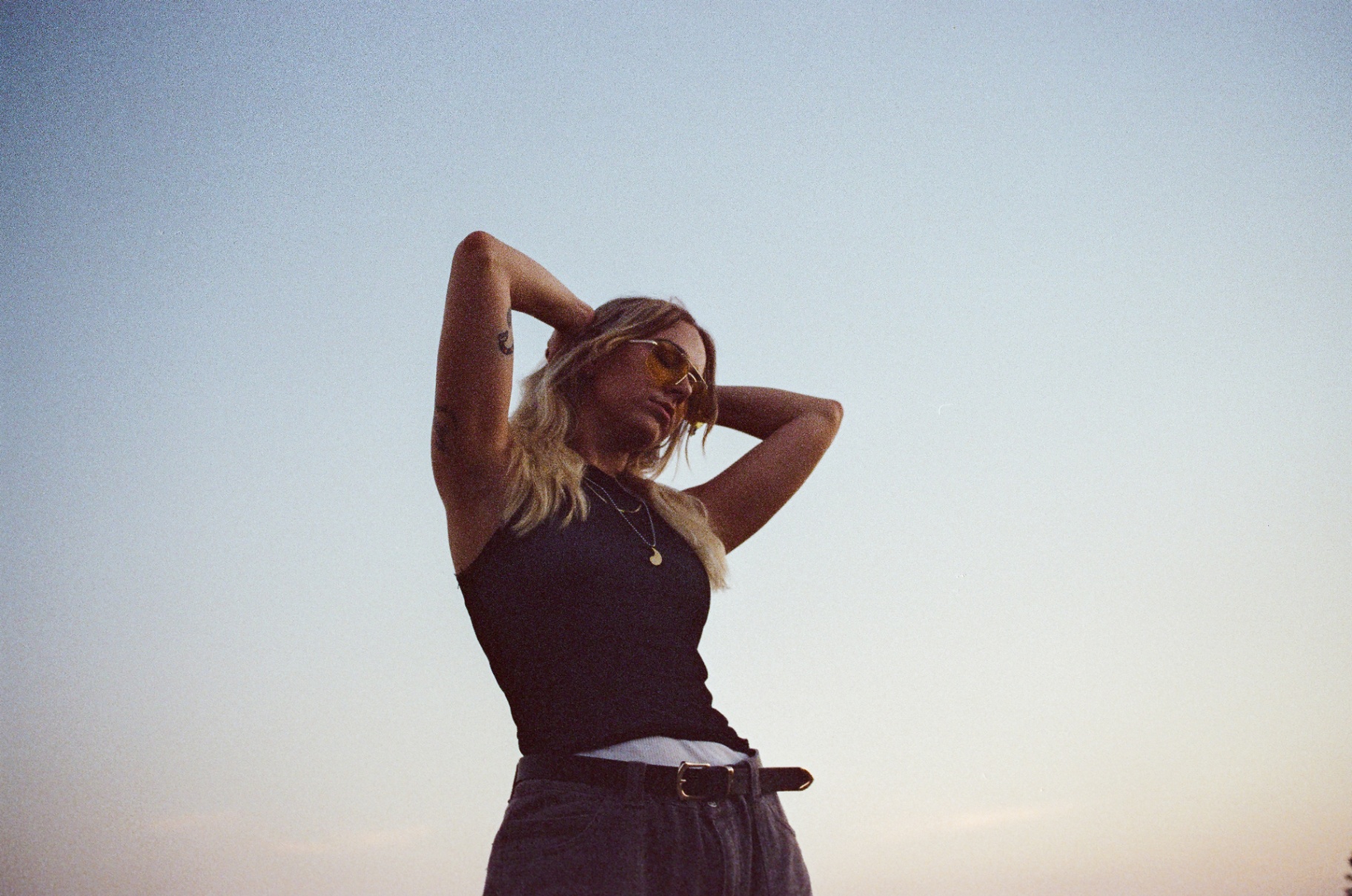 Original low-angle 35mm portrait of a woman in yellow sunglasses with her arms raised against a dusk sky