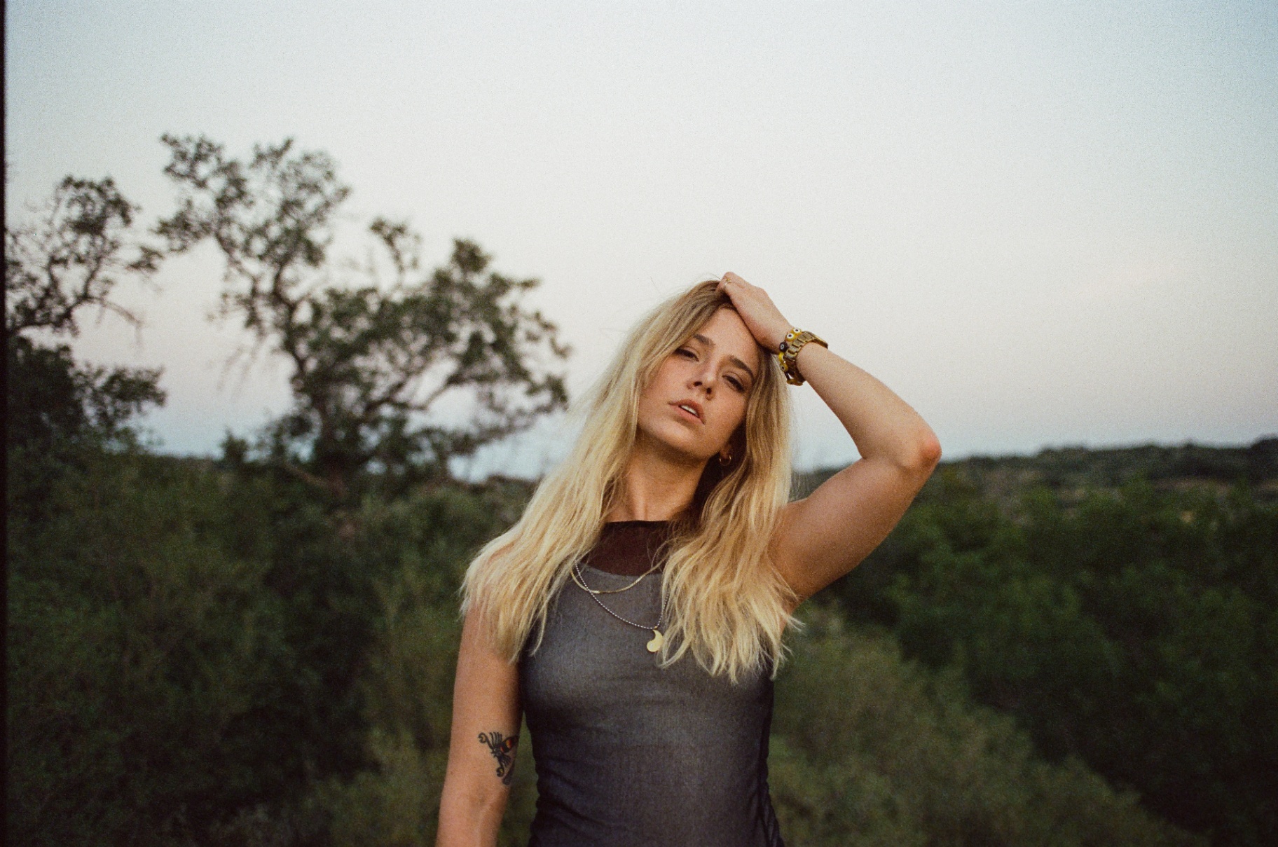 Original 35mm portrait of a blonde woman with a tattoo among green trees under a white sky